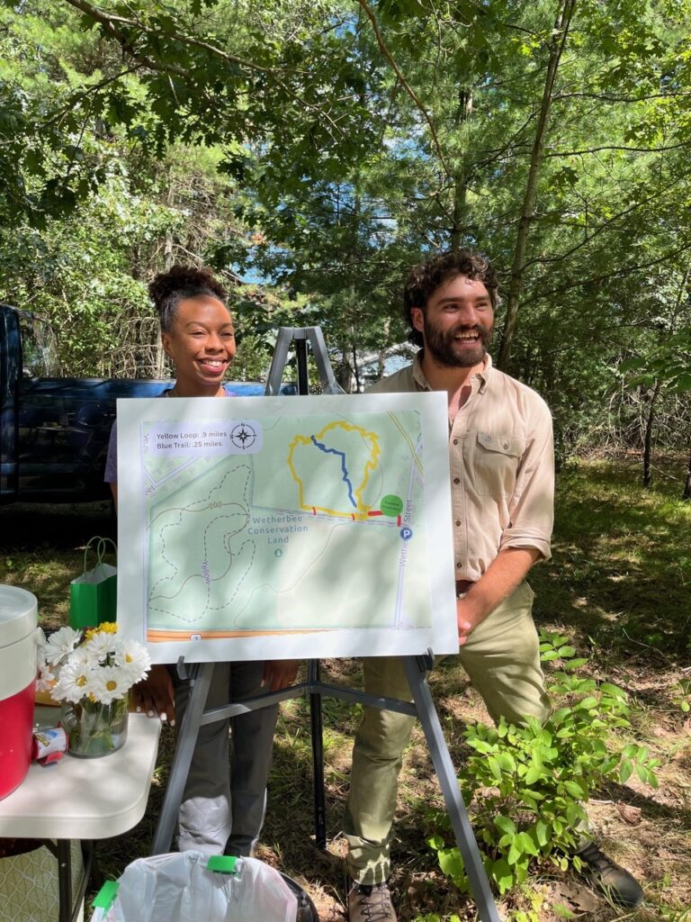 A smiling woman and man stand behind a map of the newly-purchased land (and how it connects to other town land).