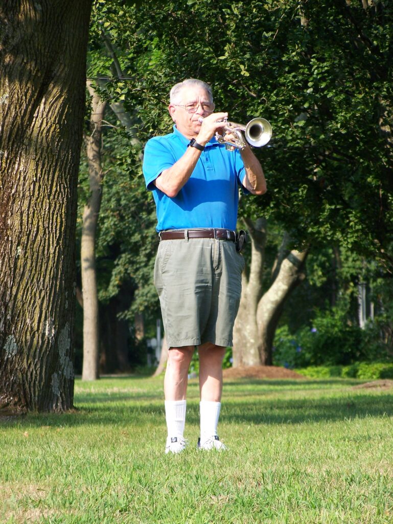 An older man, also in blue shirt and shorts plays the bugle echo from under some trees.