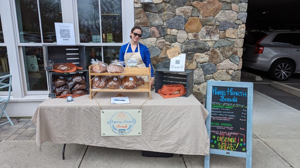 A woman stands at a table with a Harris Hearth Bread sign. The table is full of loaves and packages of English muffins.