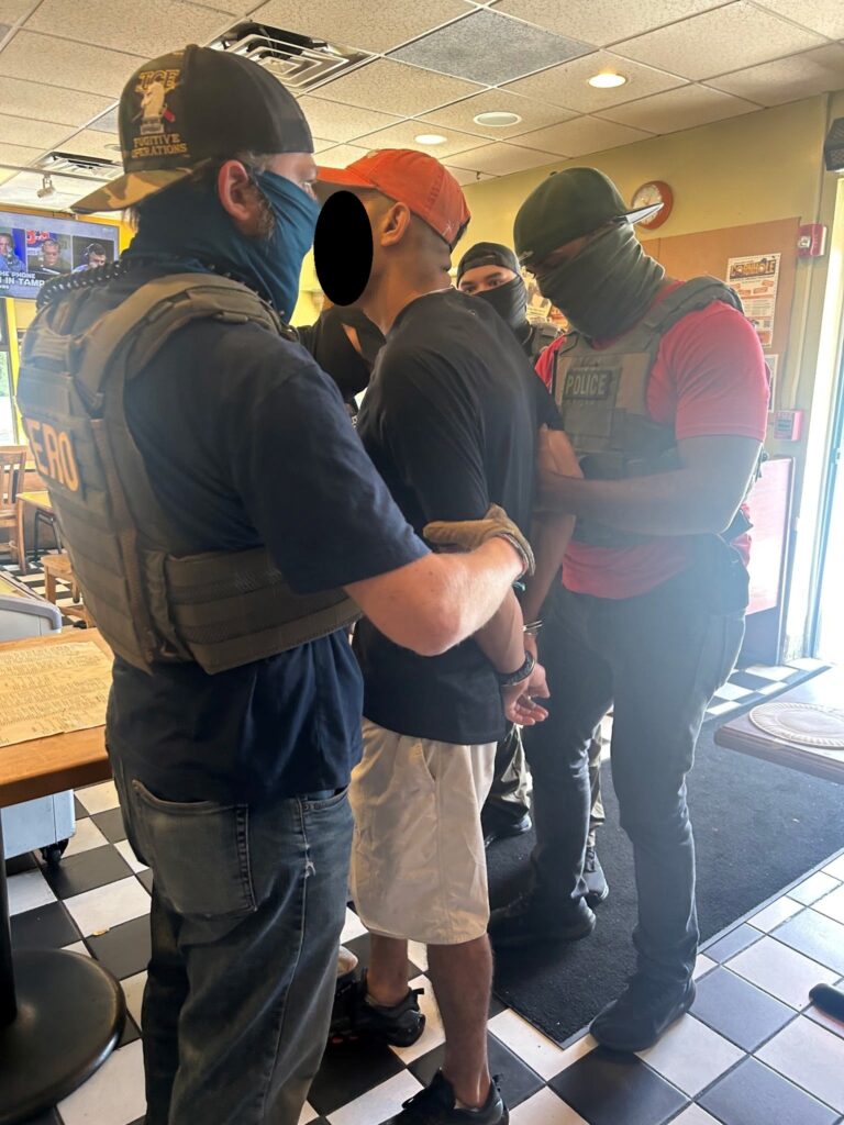 Four men wearing masks and bullet-proof vests surround and handcuff a young man inside a restaurant.