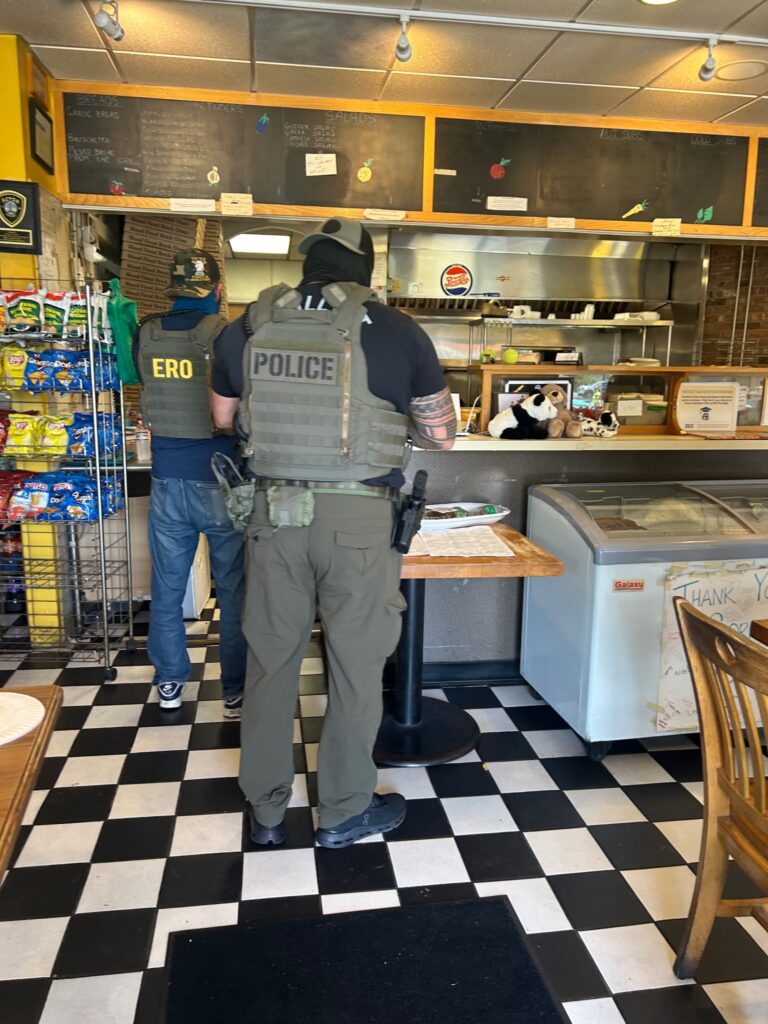 Two men wearing bullet-proof vests, face coverings, and hats stand at the counter in Sorrento's.