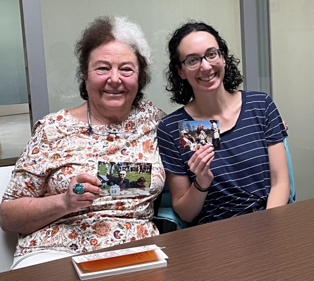Two smiling women, one with large white streak in her hair, lean into each other and hold pictures of Charlotte Sagoff.
