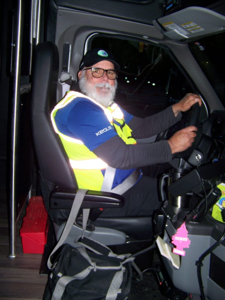 A bearded man wearing a Keolis t-shirt and a yellow vest sits at the wheel of a vehicle.