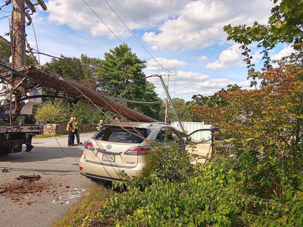 A car sits by the side of the road. A utility pole is being lifted off the car by a truck that is off to the left of the picture. There are a few people in yellow vests standing around.