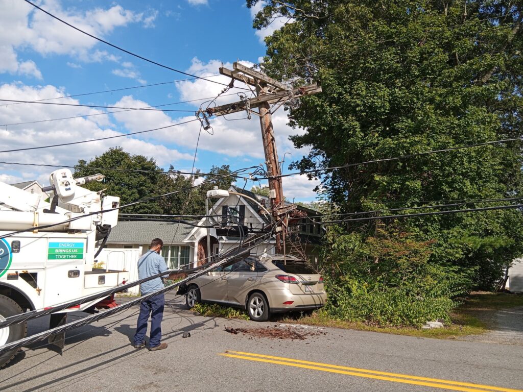 The car is still by the road. It looks like the pole is getting lefited back up (but it's a little hard to tell). The back of a utility truck is in the foreground.