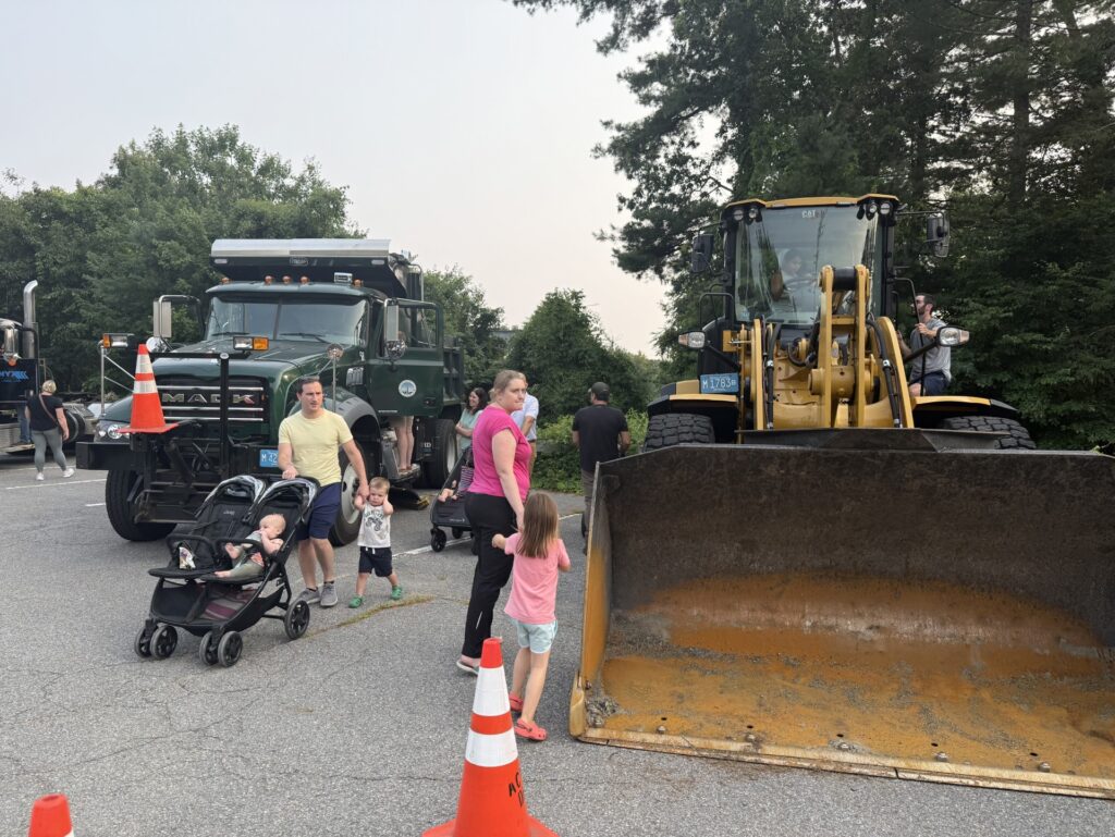 Some highway department vehicles are out on the pavement. A small boy wallks with his dad, covering his ears. Other people check out the truck and front-loader.