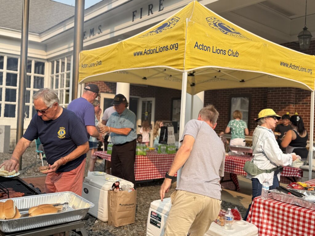 A yellow Lion's Club tent is busy with people making and serving burgers and hot dogs.