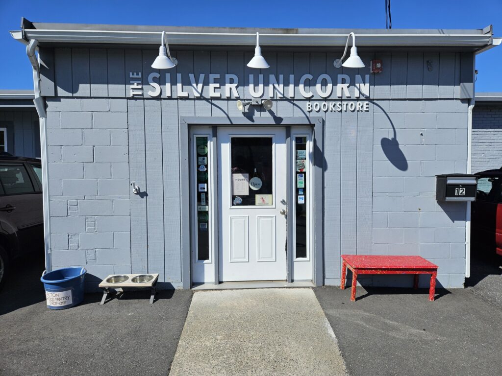 A small grey brick building with a white door. The sign says "The Silver Unicorn Bookstore." On one side of the door is a food pantry drop-off bucket and a dog water bowl. On the other side, a cheerful red wooden bench.