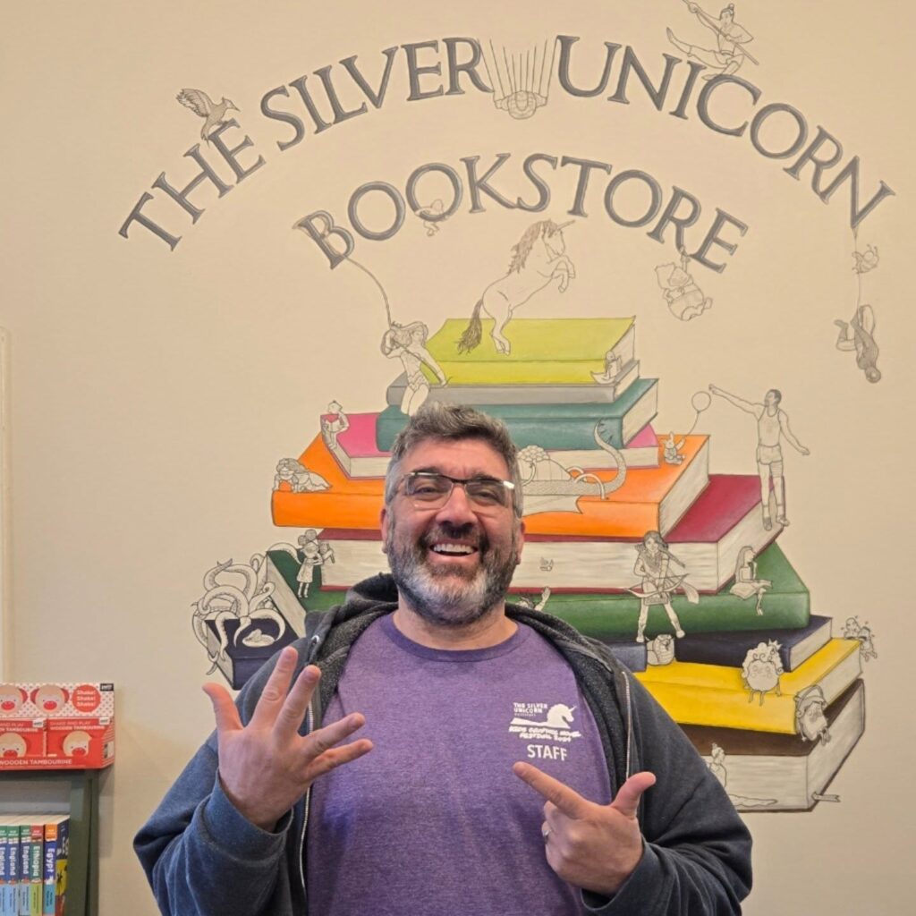 A smiling bearded man wearing a purple t-shirt counts to seven on his fingers. He stands in front of a mural with a stack of books with creatures, real and imaginary sitting, standing, climibing, and swinging from both the books and the Silver Unicorn Bookstore text that arches above.