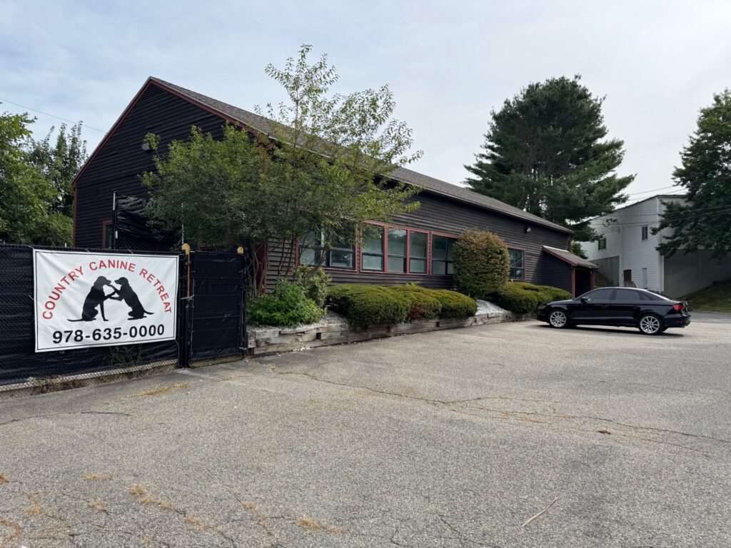 A brown building with a car parked out front. A fence has a sign on it that says Country Canine Retreat and has a silhouette of two dogs playing and a phone number.