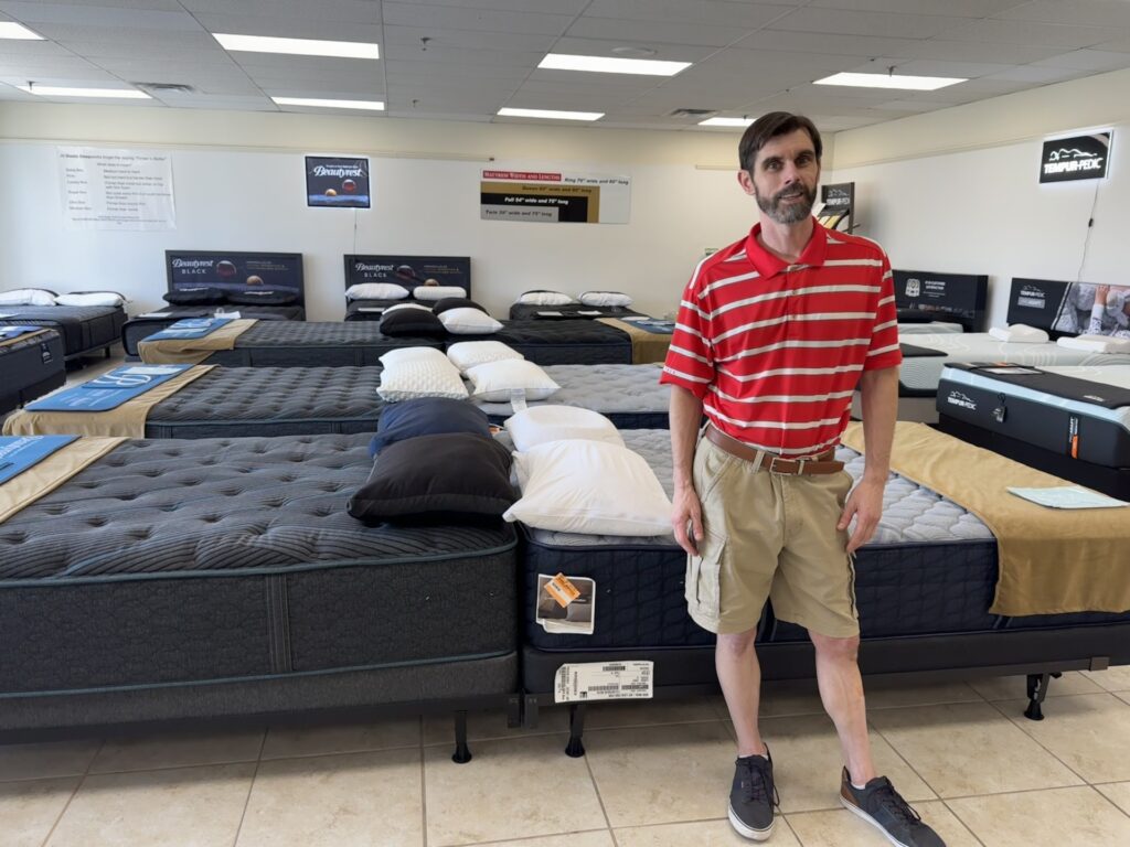 A bearded man wearing a red polo shirt and shorts stands in front of a whole line of mattresses.