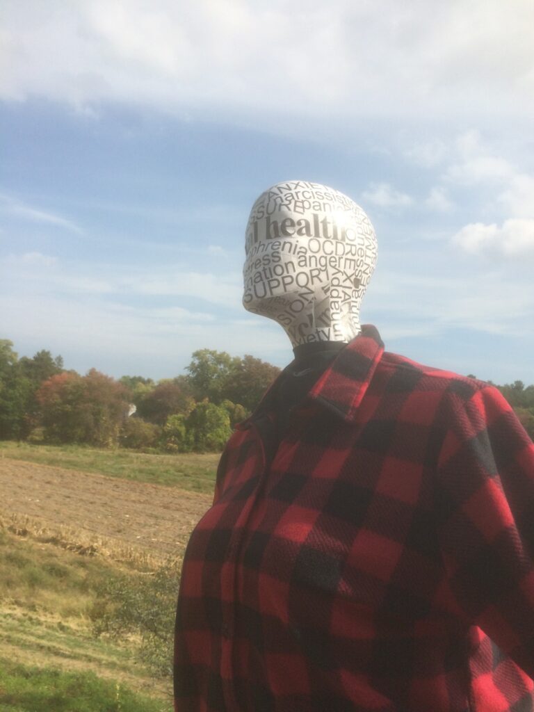The top half of a scarecrow. The head, possibly made of papier-mâché, has writing all over it -- words such as Mental Health, anger, and Support. The torso sports a red and black flannel shirt.