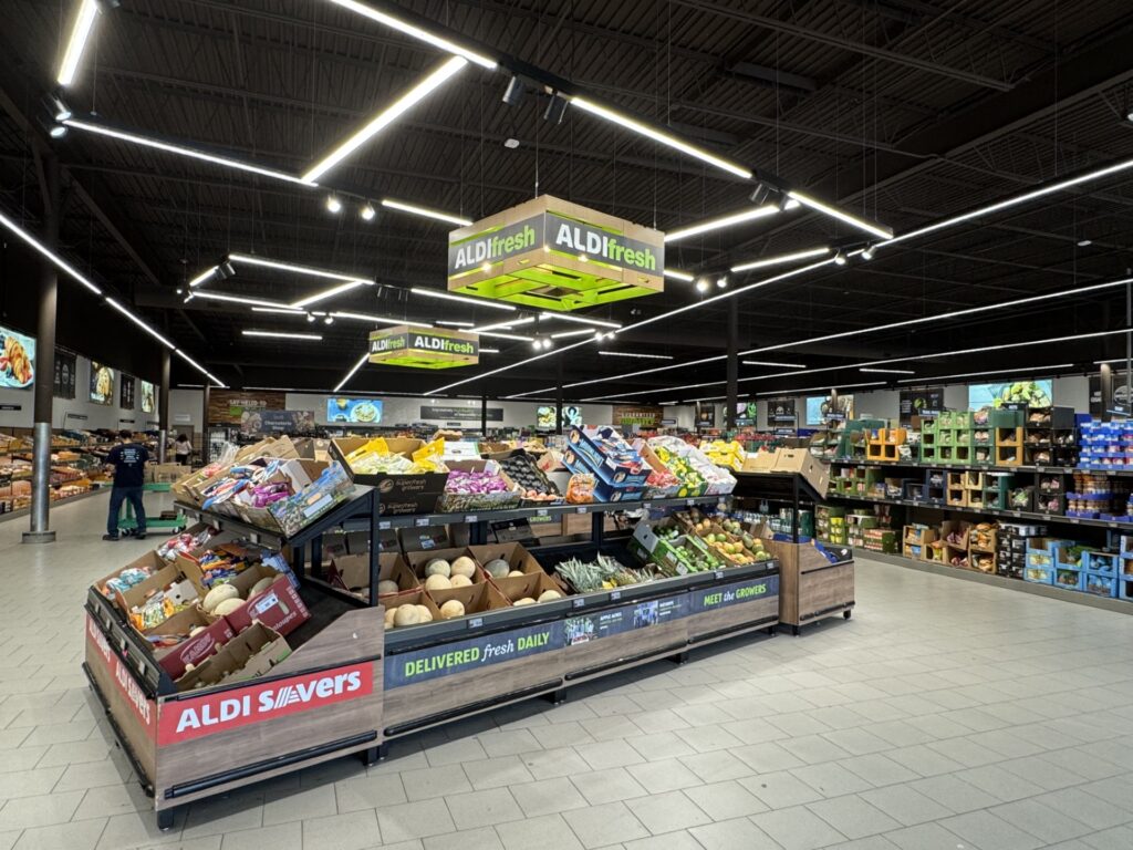 Lots of fruit piled in a large supermarket display. Other foods line additional displays on the shelves lining the room. Large, square beige tiles make up the floor around the central display.