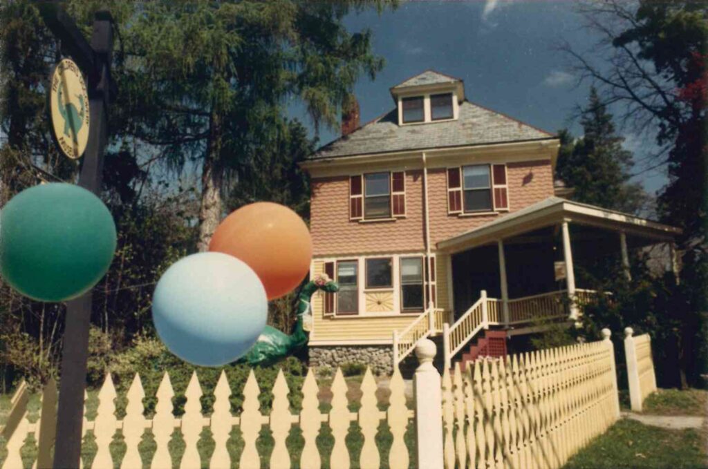 Big old square wooden house of orange and yellow color, with maroon window shutters. Porch on right side. Yellow picket fence and helium balloons in foreground.