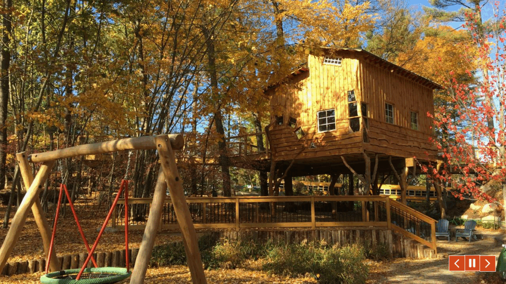 Fall colors of leaves surround a large, brown, vertical-board wooden treehouse and wooden walkways and a wooden frame above a giant round swing.