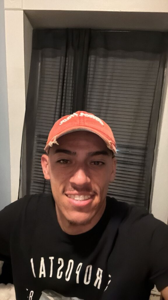A selfie of a smilling young man wearing a red cap and black t-shirt.