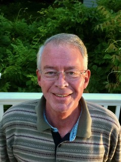 Photograph of a man with silver, short-cropped hair, glasses, a friendly smail. Grey striped shirt.