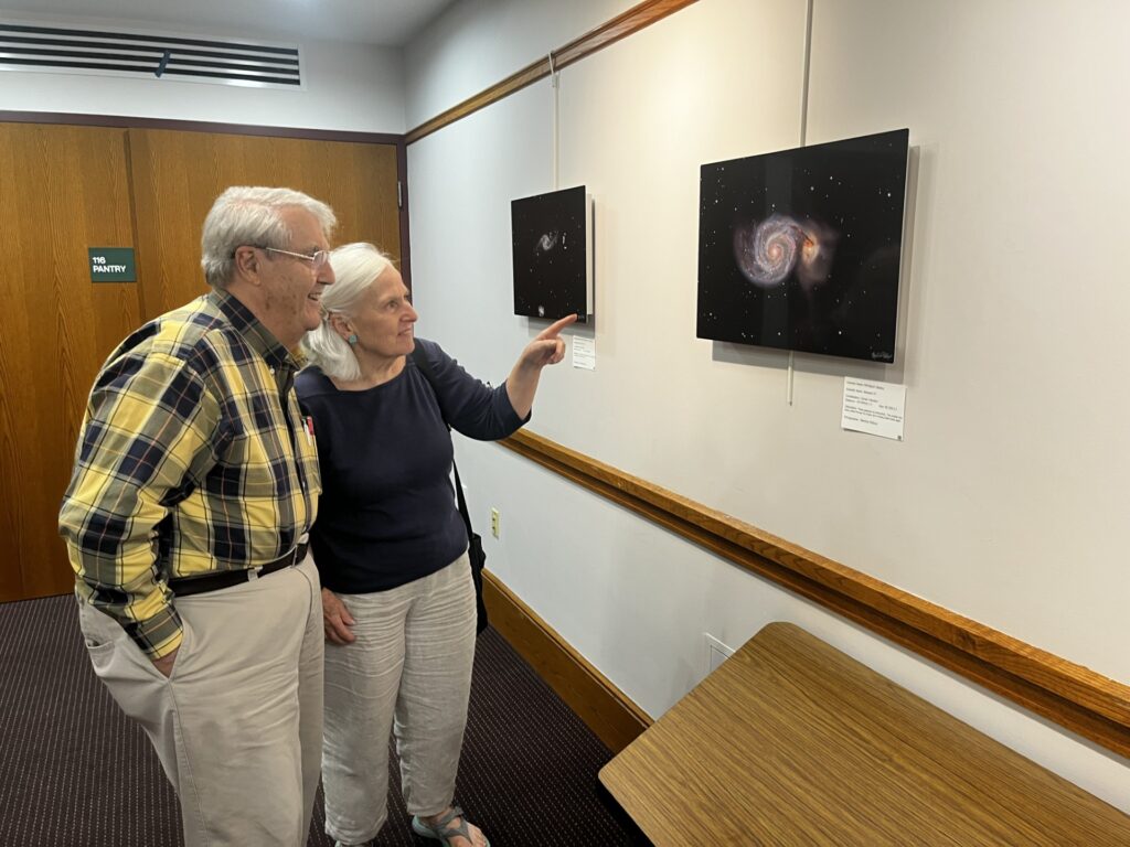 Two grey-haired people view and point to a framed photograph of black space with a colorful spiral galazy shape. Man has glasses and a yellow and black plaid flannel shirt. Woman ahas light blue earrings and a black long sleeve t shirt.