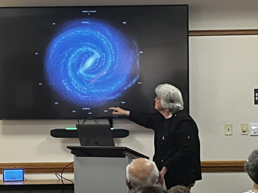 Woman with grey hair next to a podium points to a slide of a blue image of the Milky Way on a black background. In the foreground is the top of some audience members' heads.