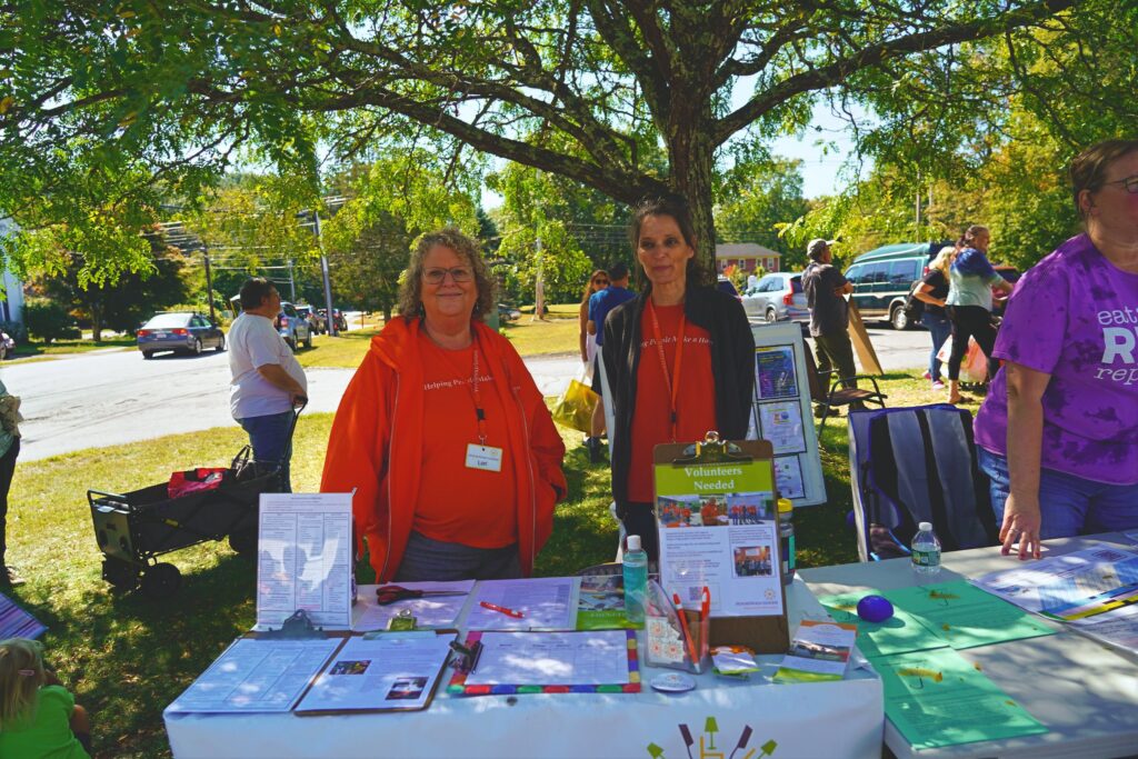 Two women wearing orange Household Goods t-shirts stand in front of a table with information about Household Goods.