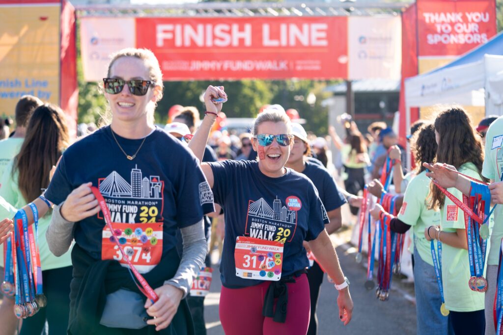 Two smiling women wearing Jimmy fund t-shirts are walking down a busy street. One of them has their fist raised. On the sidelines are volunteers handing out medals. In the background is a large sign that says "Finish Line."