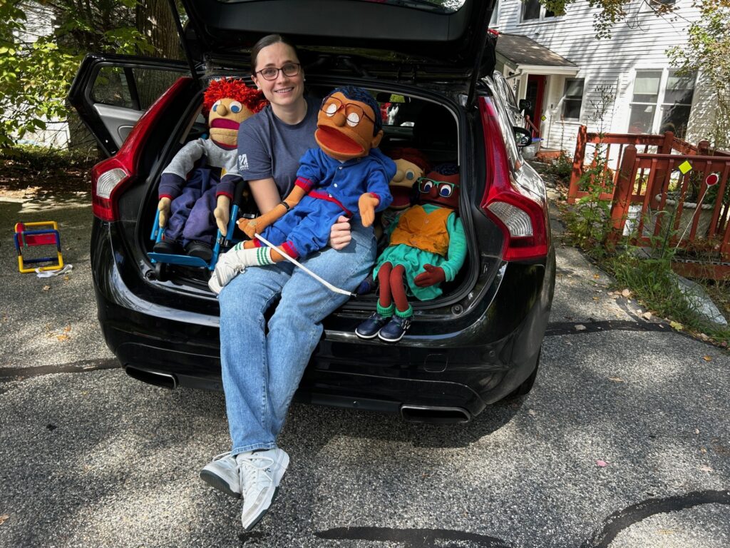 A woman sits in the back of a car with four large (roughly 3-feet tall) cloth puppets. One of the puppets sits in the woman's lap.