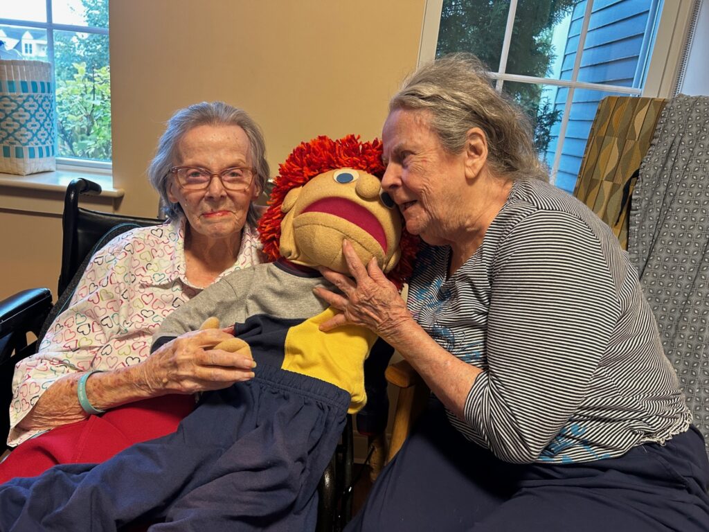 Two older women sit with one of the puppets.