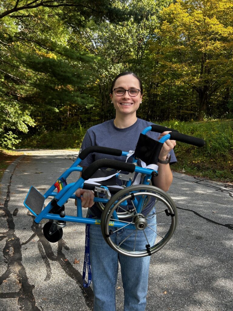A smiling woman holds a small blue wheelchair that belongs to one of the puppets.