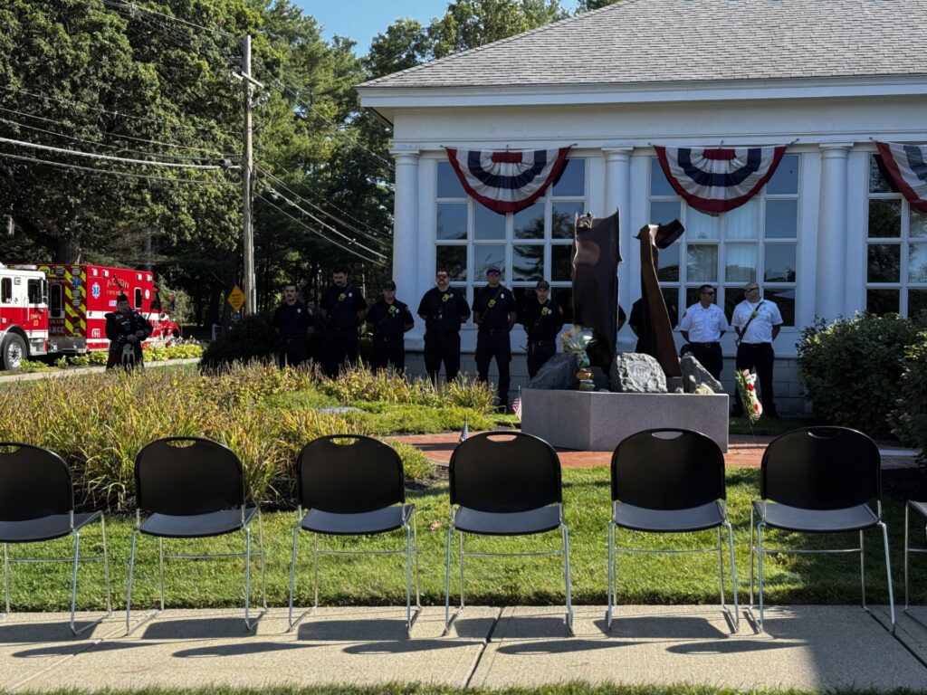 In the foreground, a group of empty seats. In the middle, the 9/11 Memorial reaches towards the sky, and in the bachground, people are lined up at ease in front of the Public Safety Building.