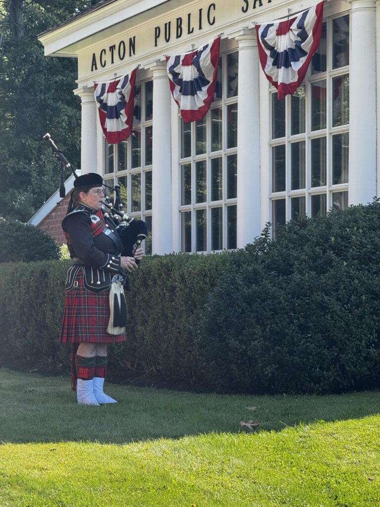 A man in formal Scottish dress (from hat to kilt to white boots, plays a bagpipe from the corner of the lwan in front of the Public Safety Building.