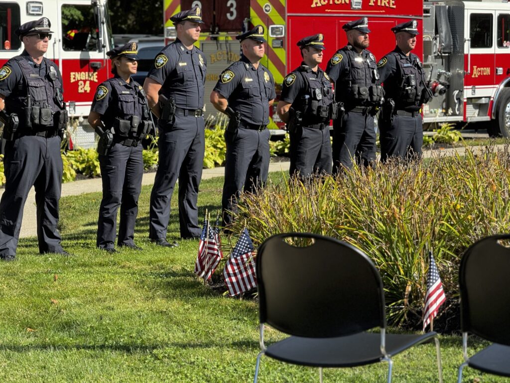 Police in uniform line up in front of some fire trucks.They are standing "at-ease," hands behind their backs.