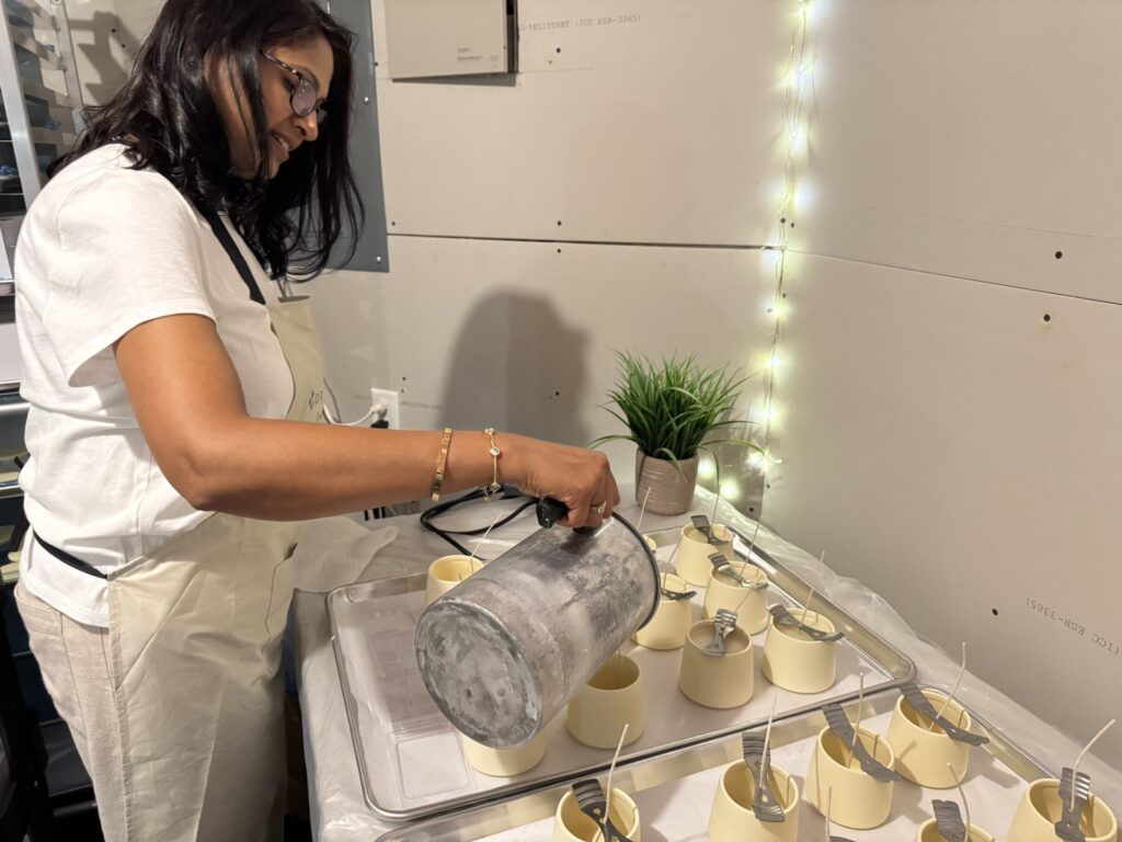 A woman pours wax from a large metal container into multiple ceramic candle holders. The candle holders have clips to hold the wicks up while the wax cools.