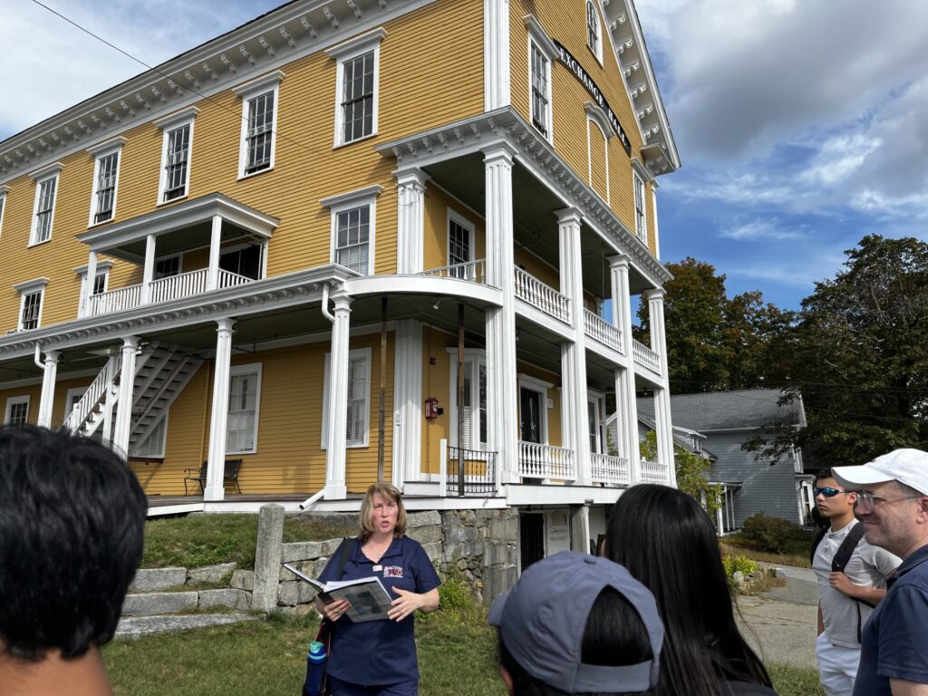 A blonde woman carrying a notebook stands in front of a large, mustard-colored building. In the foreground are the heads of people listening to her speak.