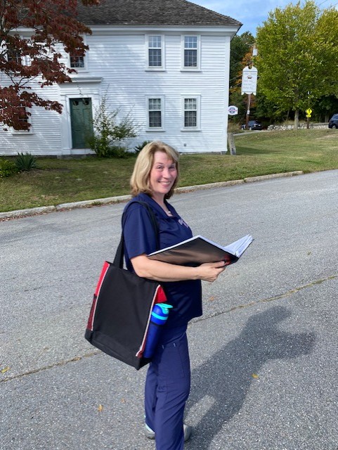 A blonde woman carrying a notebook and large tote bag stands in the street in front of a white Colonial-era tavern.