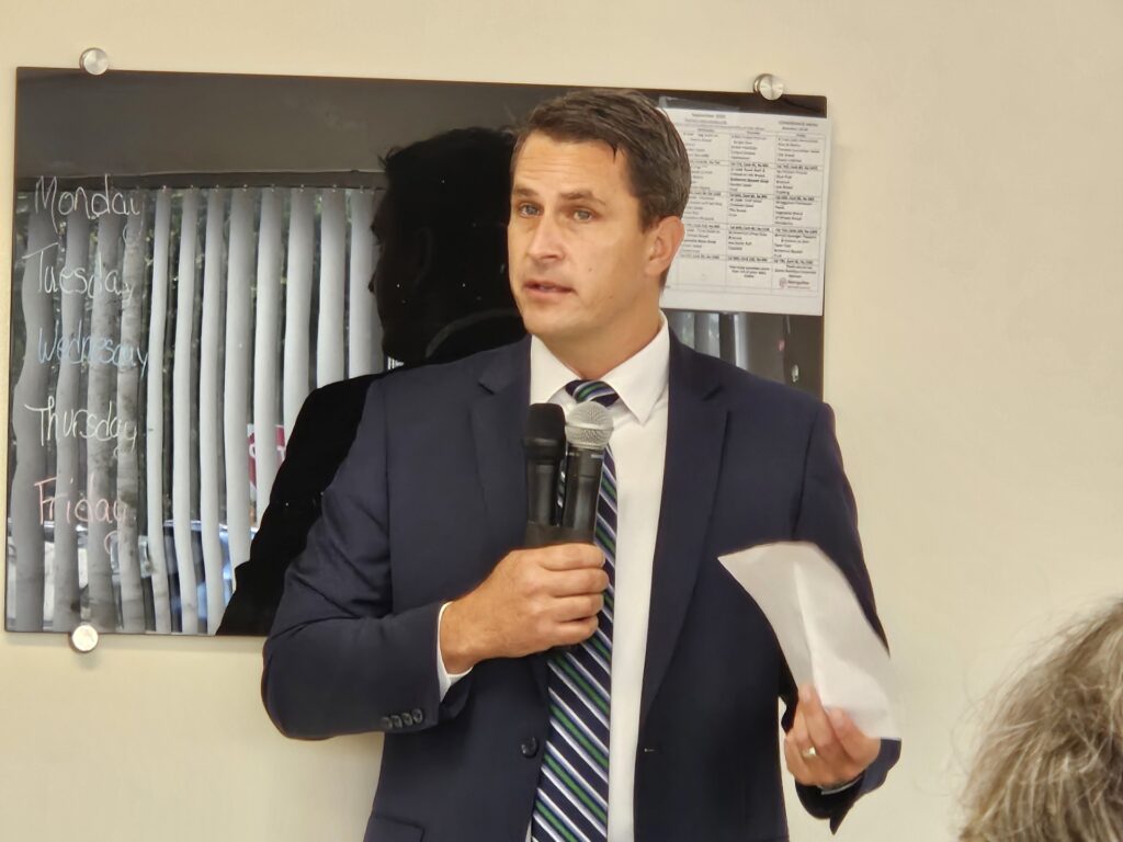 A man in a suit stands in front of a wall, speaking.