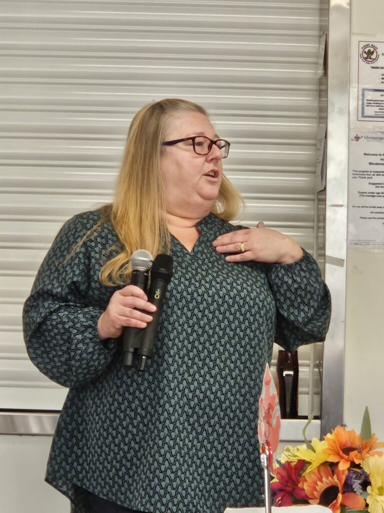 A blonde woman with glasses stands and speaks.