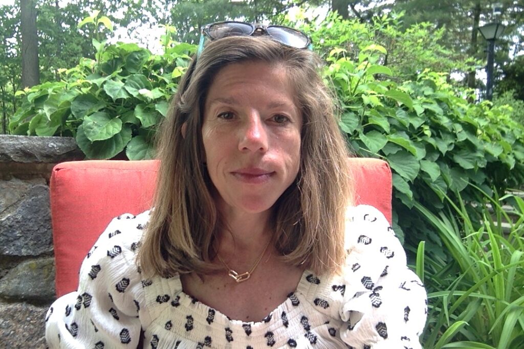 A woman with straight light brown hair and sunglasses resting on her head, seated on a coral colored cushioned chair, wearing a white and black blouse. Leaves of trees in the background.