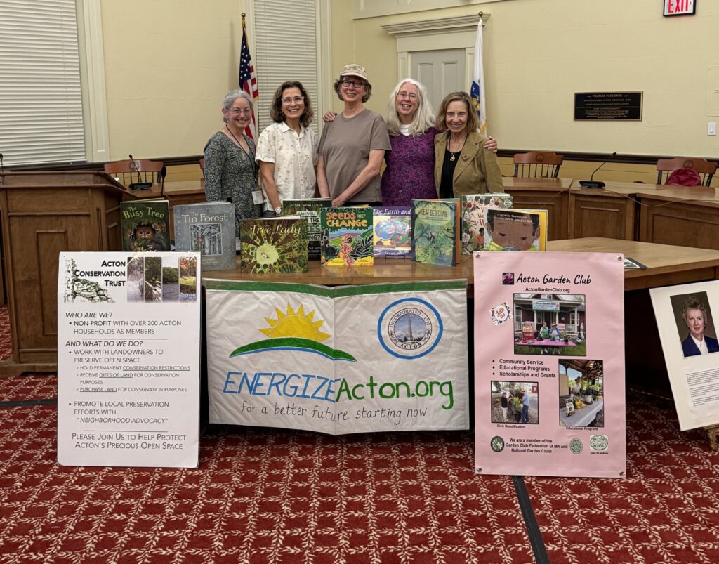 Five women stand behind a table that contains informatiom about Acton Conservation Trust, Energize Acton, and the Acton Garden Club. The table features children's books about climate and nature.