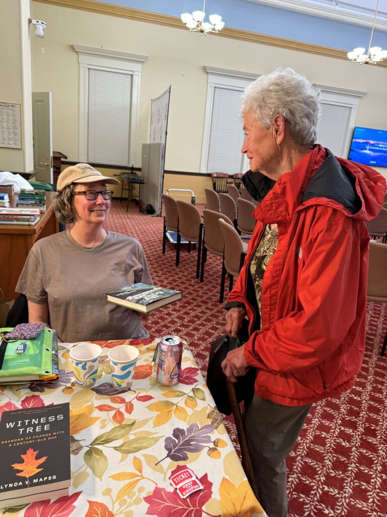 One woman, sitting at a table and holding a book is chatting with a second, older woman who is standing next to her.