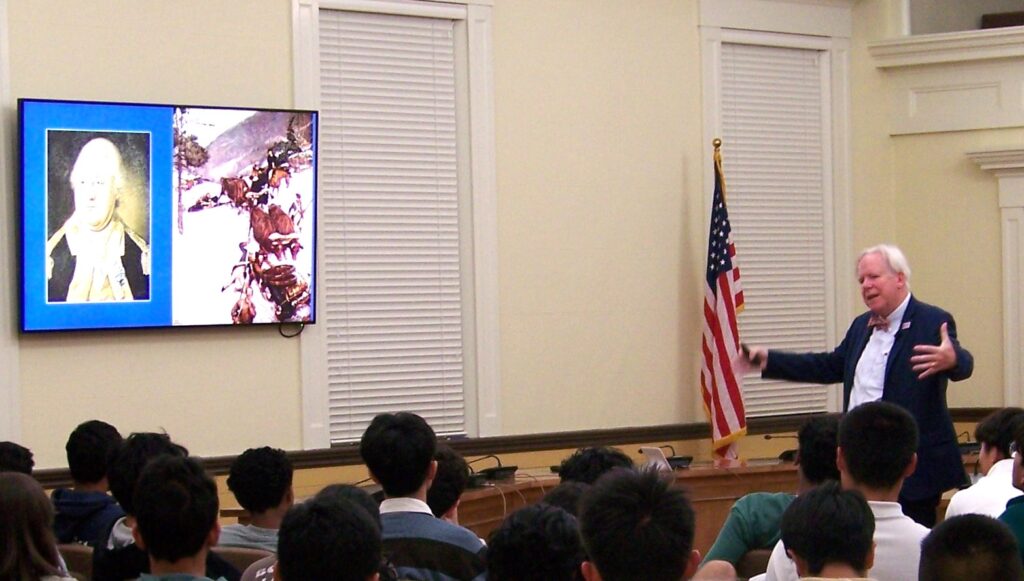 On the far right, a white-haired man wearing a bow tie gestures expansively. On the left, there's a slide show, and in the middle, a bunch of heads of people listening.