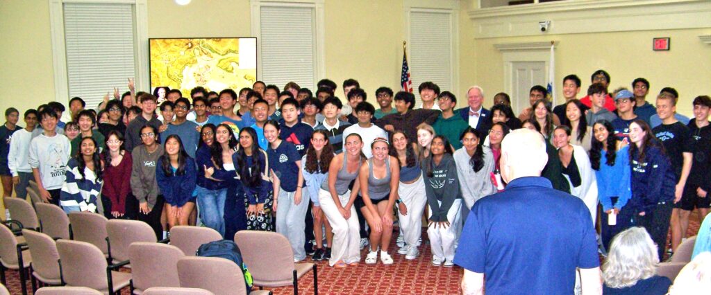 A large crowd of students poses with a white-haired man (who is at the very back). Unfortunately a man in a blue t-shirt blocks many of the students.