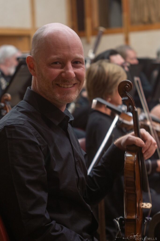 A smiling, balding man holds a violin. Other musicians are in the background.