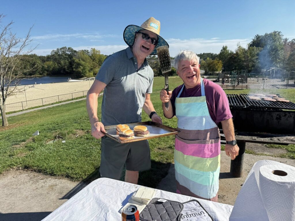 Two men, one hearing a large straw hat and holding a tray of hamburgers, the other sporting a striped apron and a large spatula, stand in front of an outdoor grill on a sunny day,