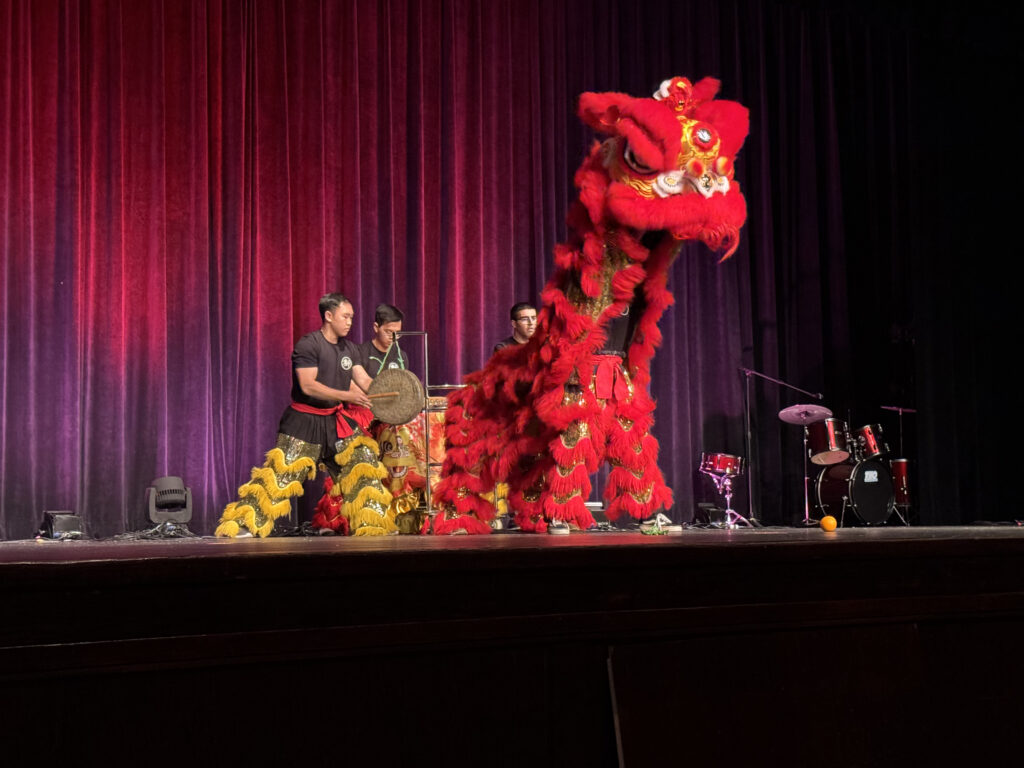 A very large red dragon towers over three performers. One performer is banging a drum and wearing an outfit with yellow fringed leggings.
