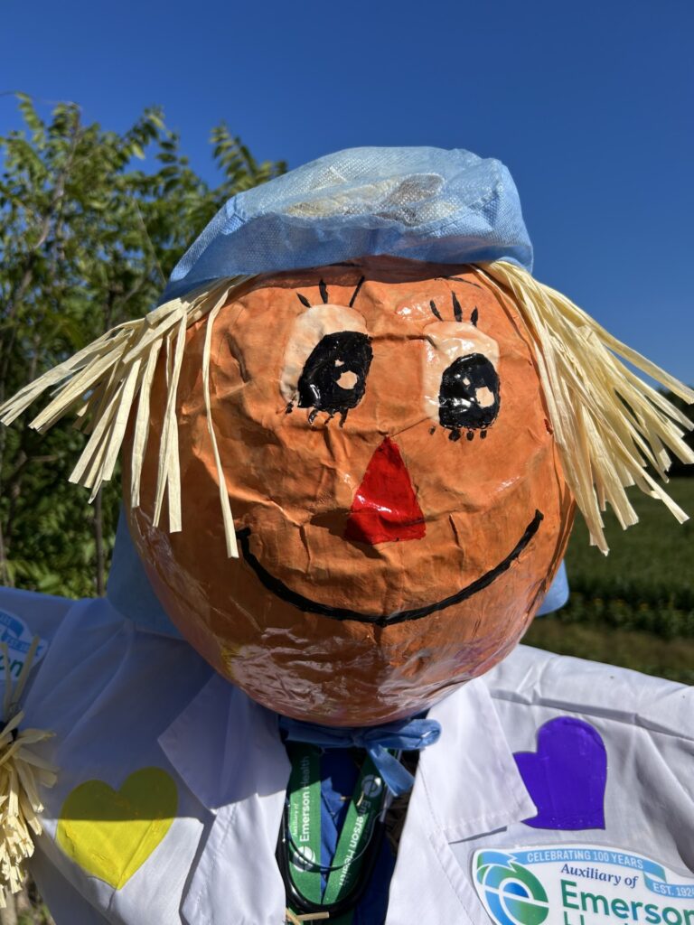 This scarecrow has a papier-mache headand wears a medical jacket and blue headcover.