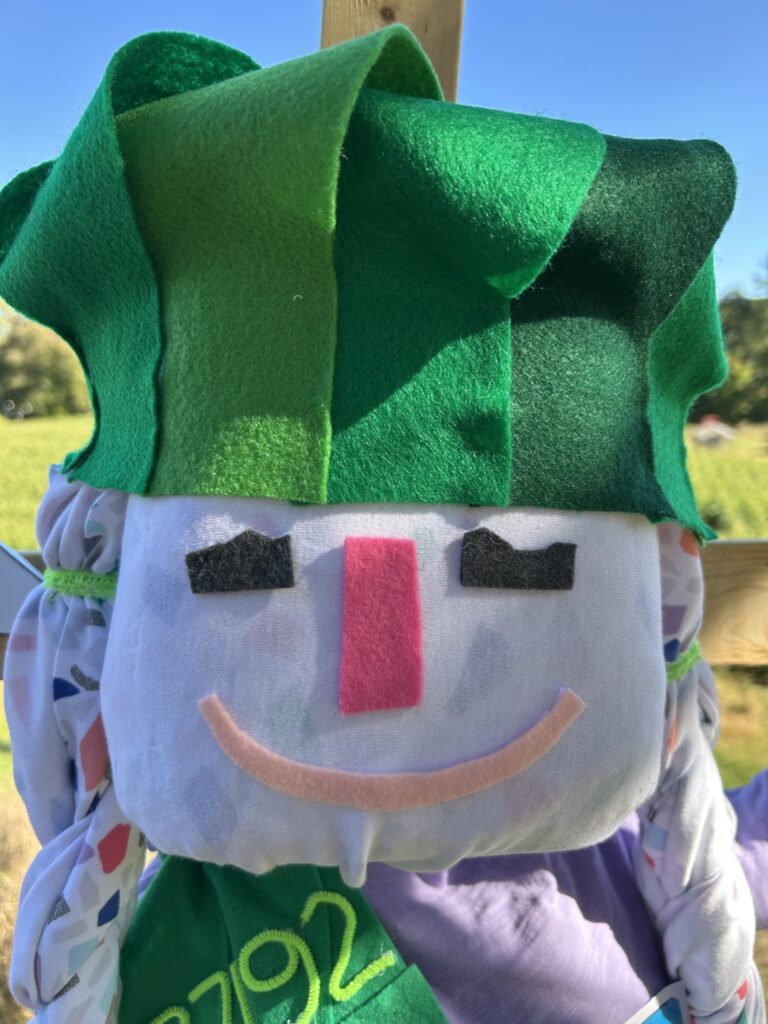 A smiling scarecrow with a felt head and a hat also made of green felt strips.