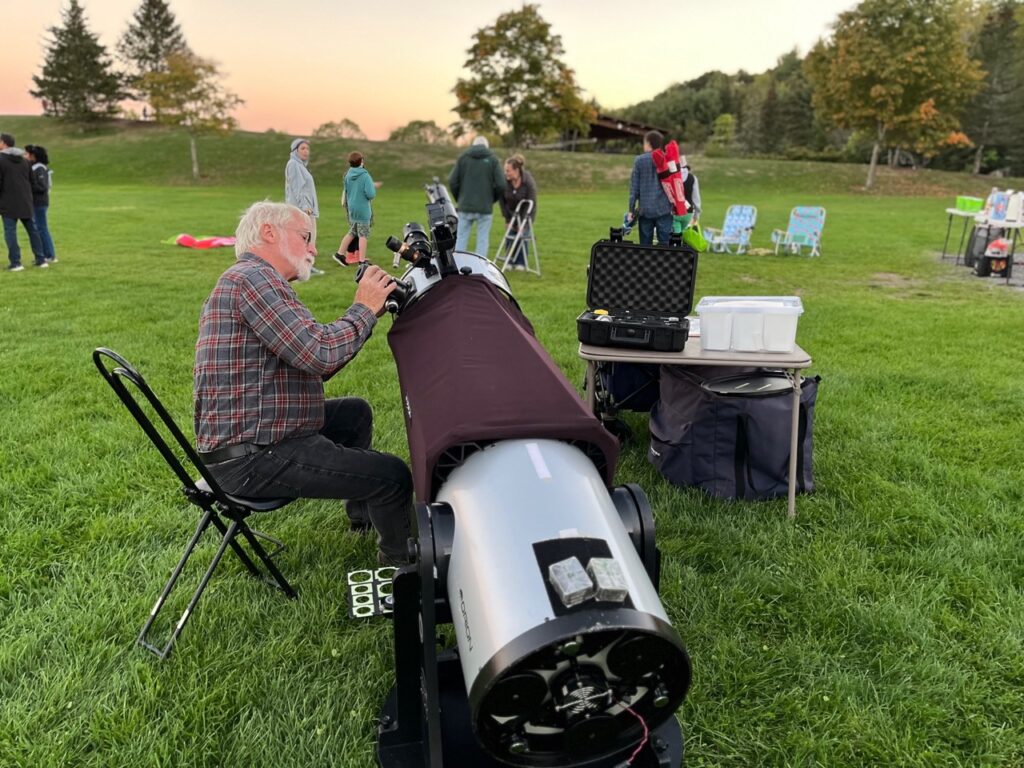 A seated man adjusts a large (maybe 5 feet long and a foot in diameter) telescope. Other people are walking around in the background.