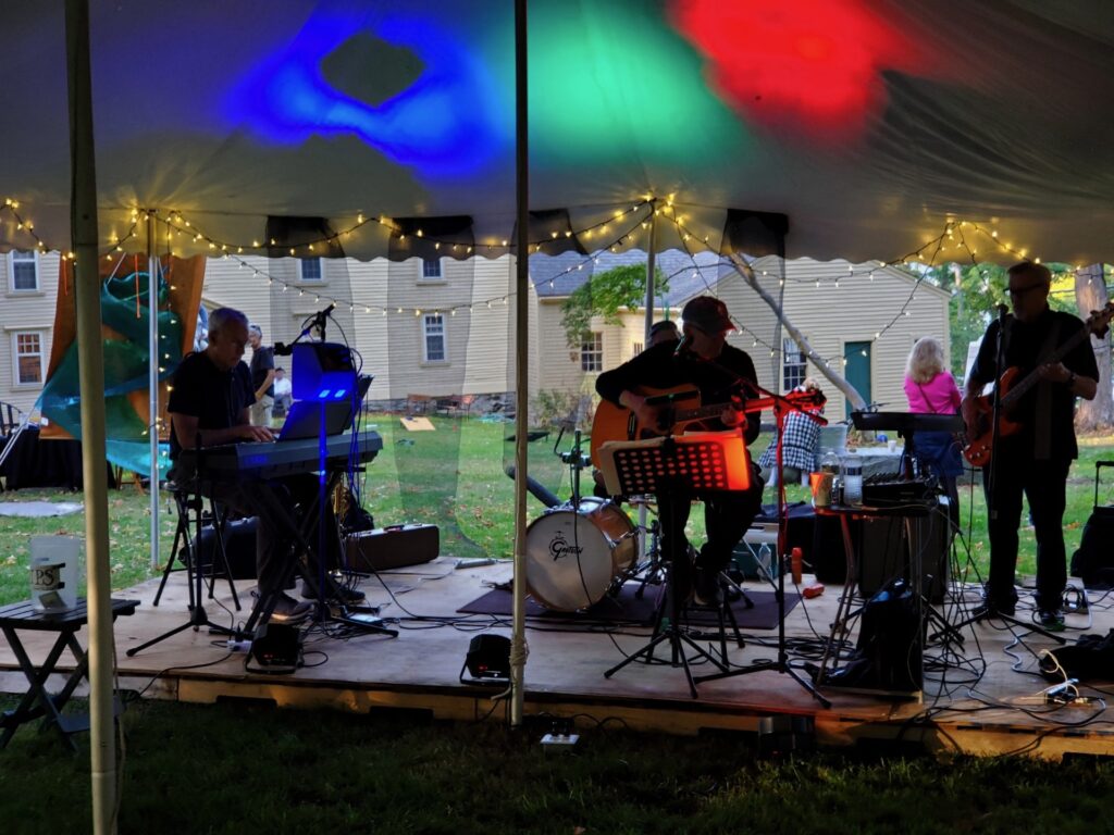 A four-piece band plays under a tent with multi-colored lights on the ceiling.