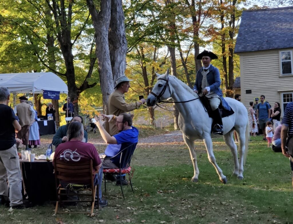 A rider on a white horse stands in front of the yellow Faulkner House. There are people sitting around chatting and people playing games at the edges of the image.