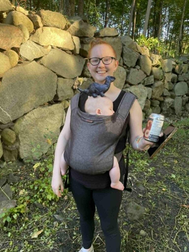 A smiling woman carries a sleeping baby in a front carrier. A small diplodocus sits on the baby's head. In the background is a stone-lined wall (a pit, actually) that is the foundation of a 19th century barn.
